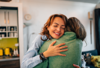 Two friends hugging for 20 seconds at home, smiling with eyes closed — the science-backed benefits of hugging