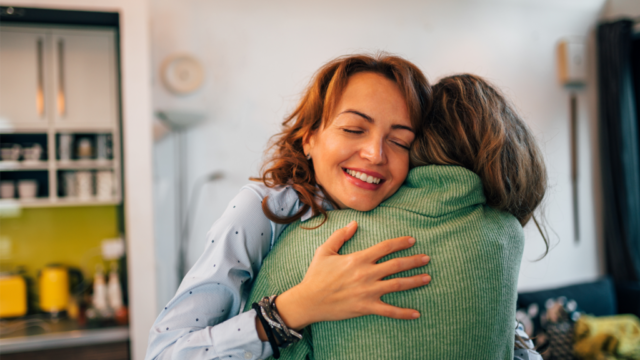 Two friends hugging for 20 seconds at home, smiling with eyes closed — the science-backed benefits of hugging